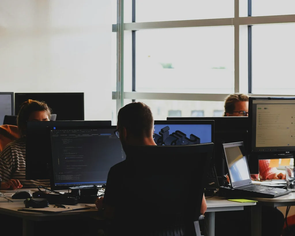 Three people are working at computer desks in a bright office. The atmosphere is focused and collaborative, with screens displaying code and designs.