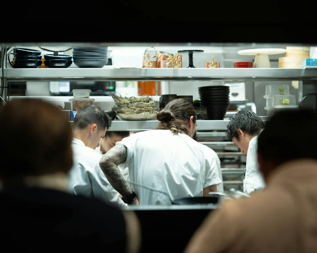 Chefs in a professional kitchen work intently, wearing white uniforms. Shelves hold bowls and kitchen tools, creating a focused, busy atmosphere.