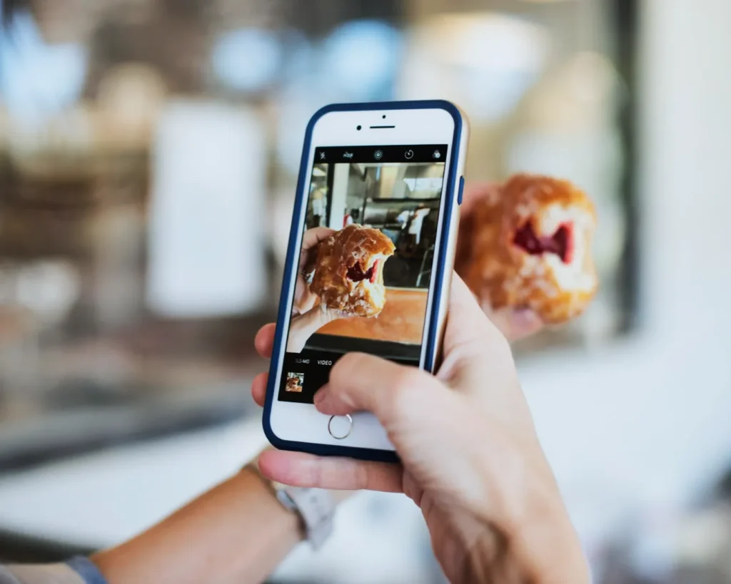 A person holds a smartphone capturing a photo of a jelly-filled donut in their other hand. The setting appears to be a bright cafe or bakery.