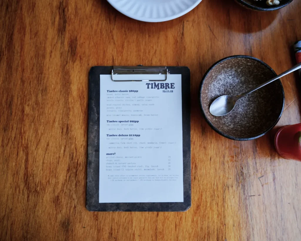 A clipboard with a menu titled "Timbre" lies on a wooden table. Nearby, there's an empty brown bowl with a spoon and a partial view of a red bottle. The scene is casual and inviting.