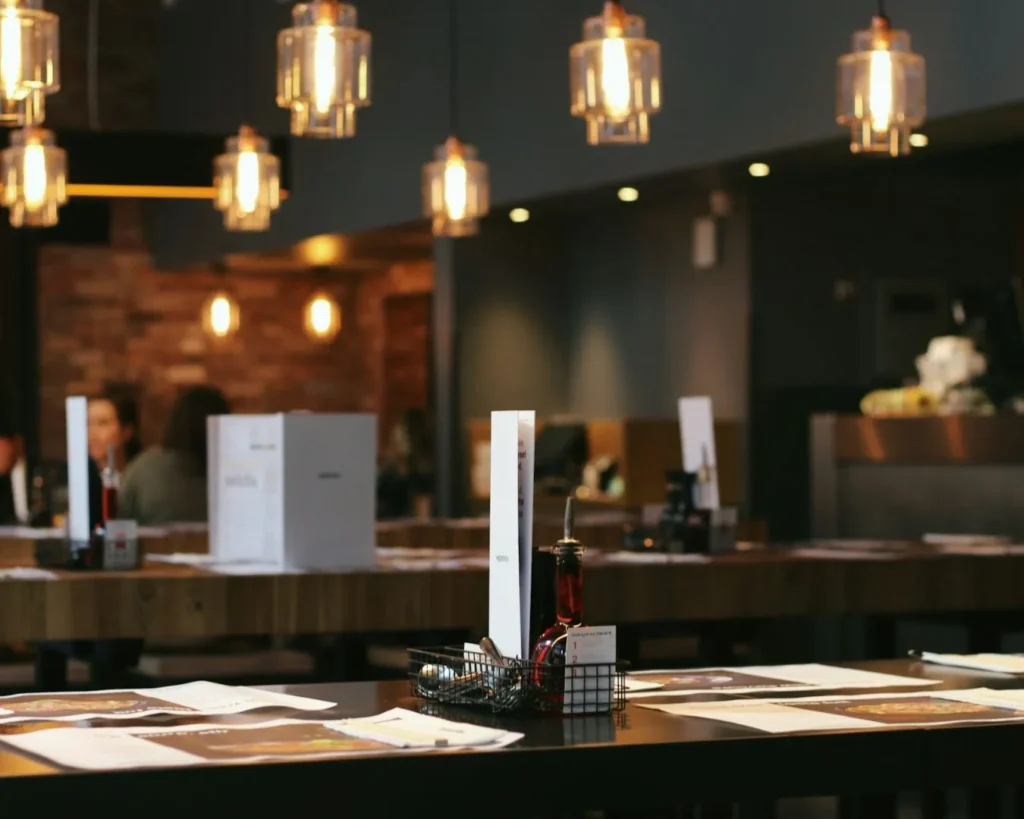 Cozy restaurant interior with wooden tables and pendant lights. Menus, condiment bottles on tables. Warm ambiance with blurred patrons and brick wall in background.