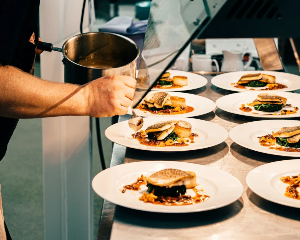 A chef artfully plates multiple dishes with fish, greens, and a rich sauce on white plates, conveying a sense of precision and culinary elegance.