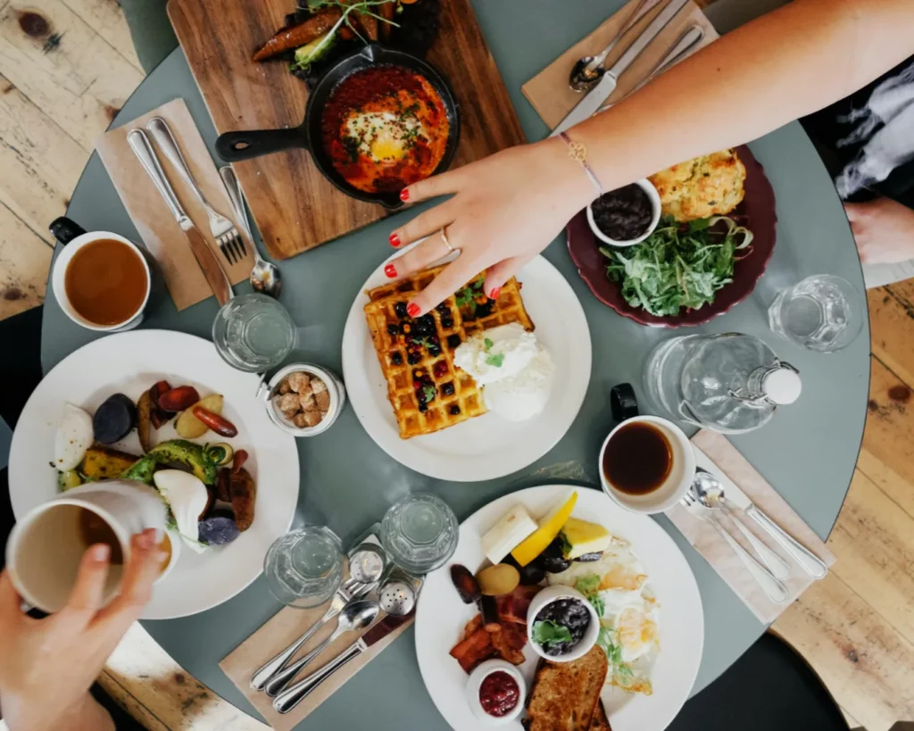 A table with diverse brunch dishes including waffles with cream and fruit, eggs, bacon, salad, and coffee. Two hands reach for the food, suggesting a lively, social meal.