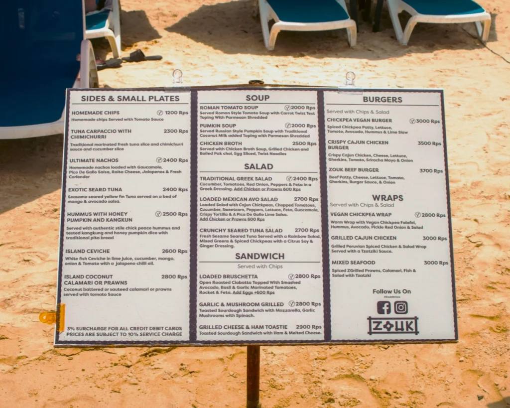 Restaurant menu on a sandy beach with blue and white lounge chairs. Categories include sides, soups, salads, sandwiches, burgers, and wraps.