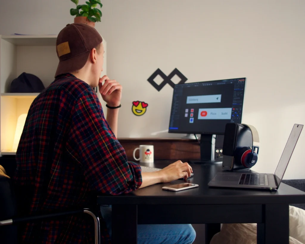 A person in a plaid shirt and cap works at a desk with a computer and laptop, surrounded by a mug, headphones, and a plant, conveying focus and creativity.