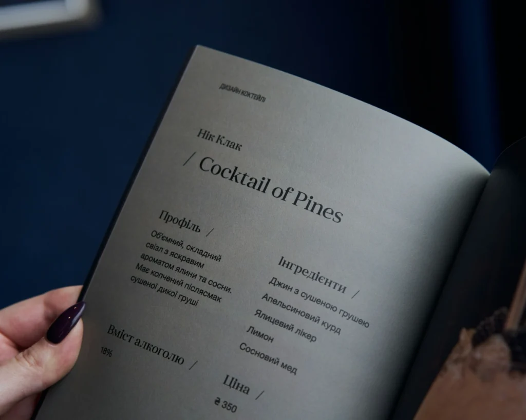 Close-up of a menu page featuring a cocktail named "Cocktail of Pines" with ingredients and details in a modern, stylish layout. A hand with dark nail polish holds the page, setting a sophisticated tone.
