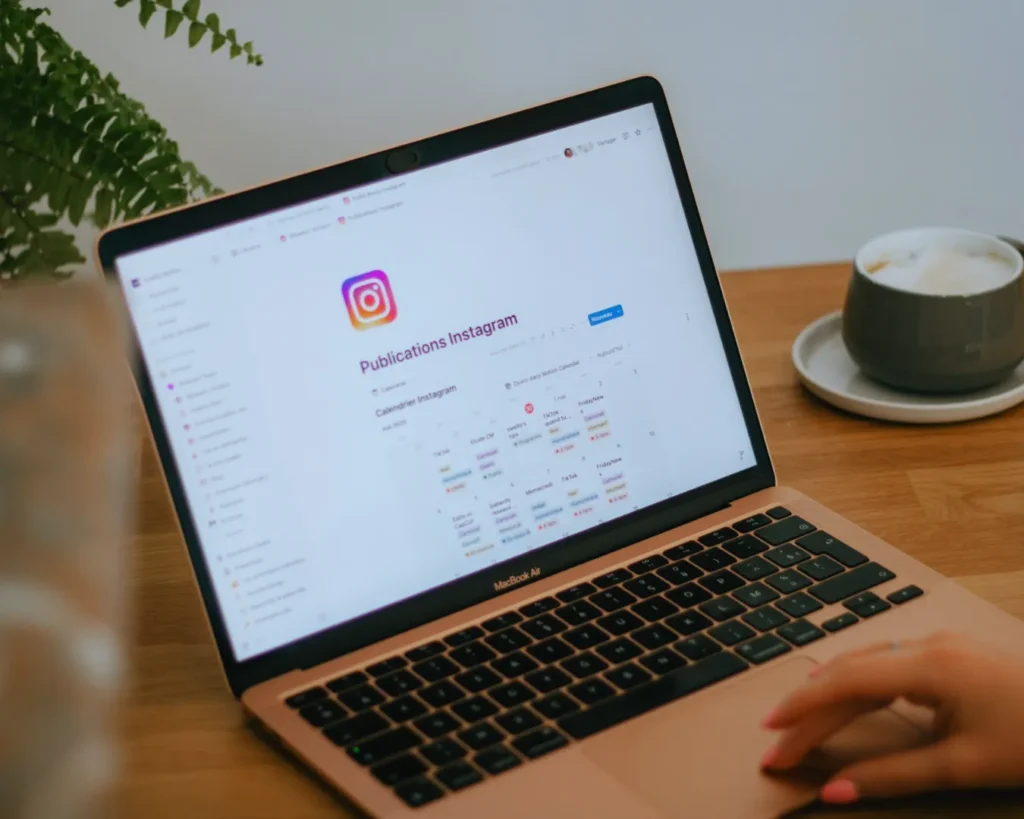 A person uses a MacBook Air displaying an Instagram content calendar. The setting is a cozy workspace with a potted fern and a cup of coffee.
