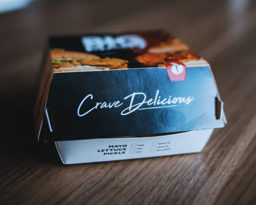 Closed fast-food burger box on a wooden table, labeled "Crave Delicious," with checkboxes for mayo, lettuce, and pickle preferences.