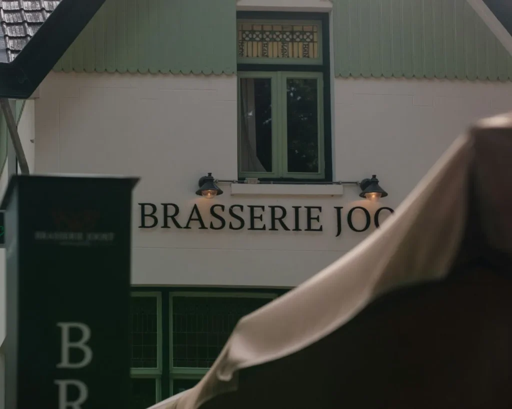 Facade of a quaint building labeled "Brasserie Joost," featuring green windows and decorative lights under a soft, overcast sky, evoking a charming ambiance.