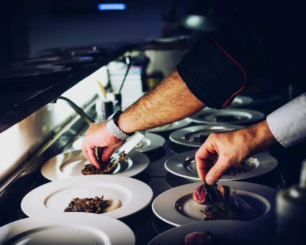 Chef's hands carefully plate gourmet dishes on a counter, showcasing culinary precision and focus. The atmosphere is professional and refined.