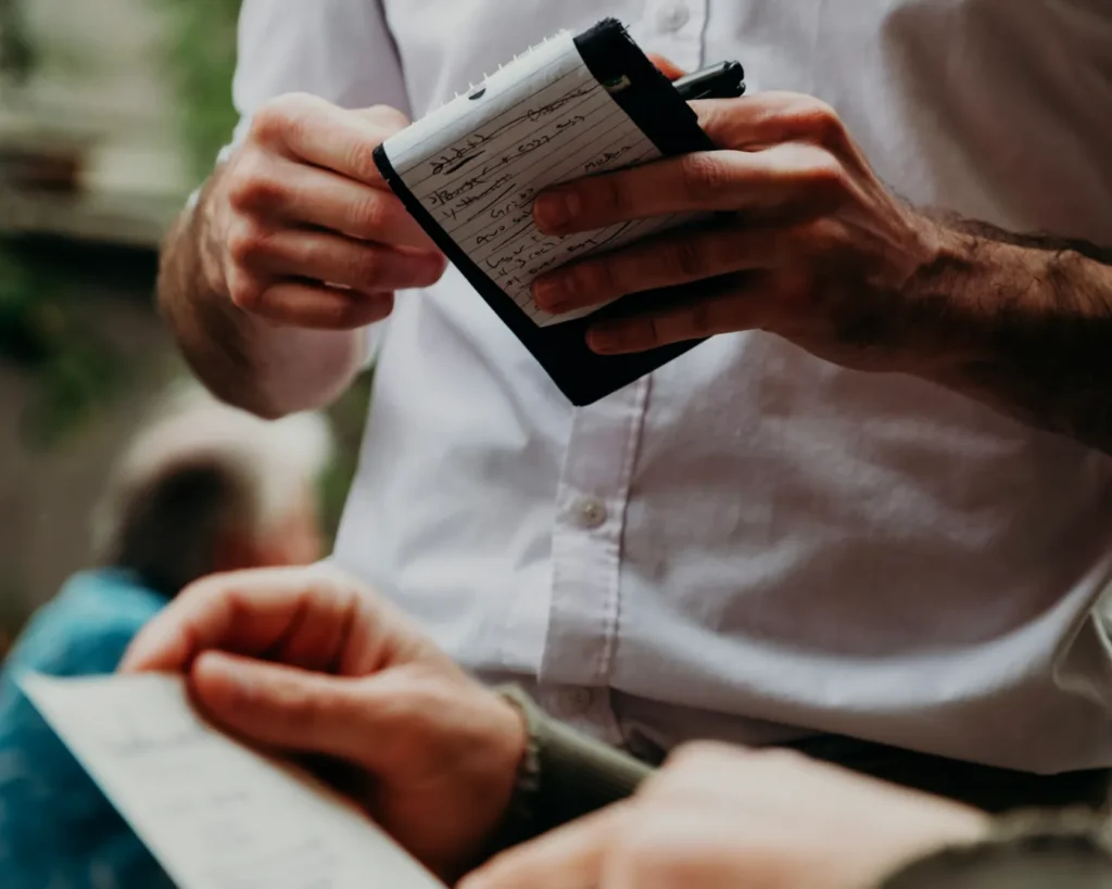 A waiter in a white shirt holds a notepad, taking an order from a customer holding a menu. The setting appears casual and attentive.