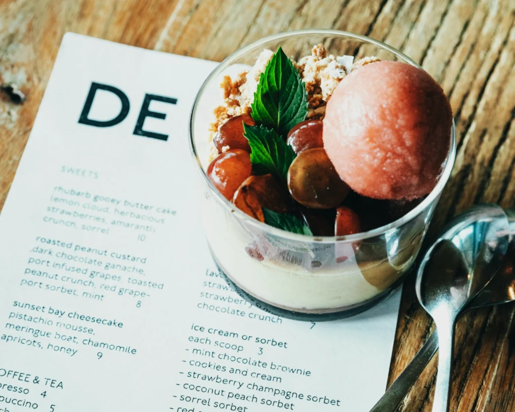 A glass dessert cup on a wooden table, containing grapes, a pink sorbet scoop, and mint leaves, sits on an open dessert menu. A spoon rests beside it.