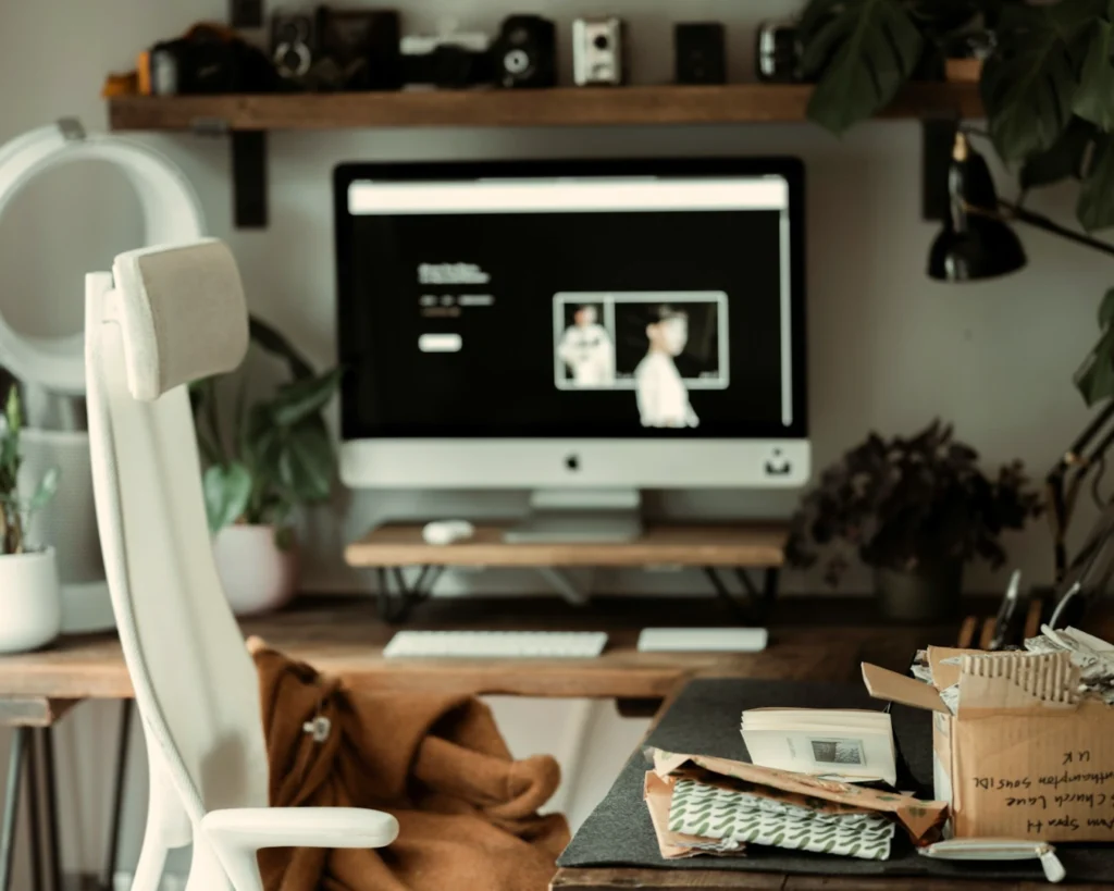 Cozy home office with a modern computer, white chair, and plants. A shelf with vintage cameras is above, while an open package lies on the desk.