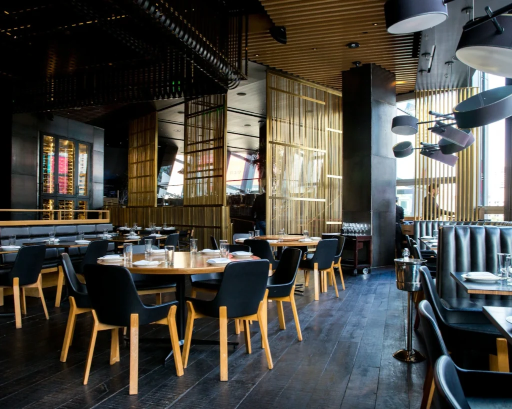 Modern restaurant interior with black cushioned chairs, wooden tables, and ambient lighting. Gold vertical accents give a sleek, elegant atmosphere.