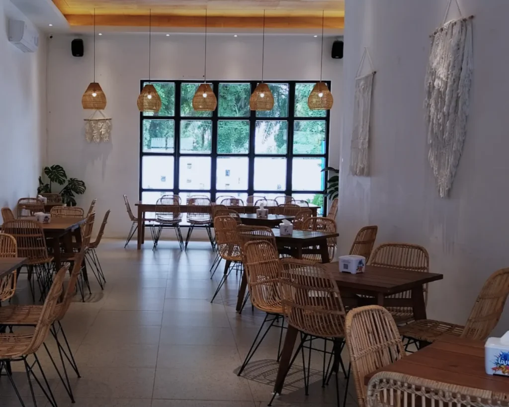 Bright, airy cafe with rattan chairs and wooden tables. Large windows, hanging wicker lights, and wall art create a relaxed, natural vibe.