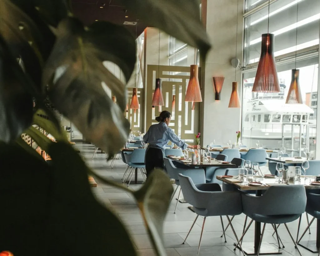 A modern restaurant interior with blue chairs and wooden tables, elegantly set. A person arranges a table, beneath warm pendant lights, near large windows.