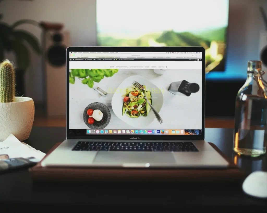 Laptop on a wooden desk displaying a website with a salad image. Nearby are a cactus plant, a water bottle, and a blurred TV in the background. Cozy atmosphere.