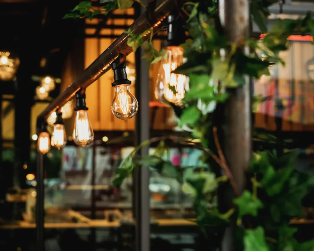 String lights with vintage bulbs hang from a rustic metal fixture surrounded by lush green ivy, creating a warm, cozy ambiance in an outdoor setting.