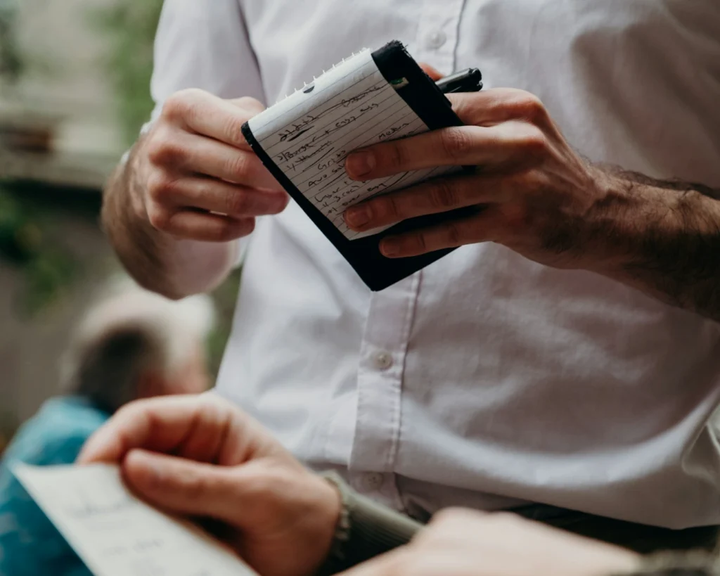 A waiter in a white shirt holds a notepad and pen, taking an order from a customer in a restaurant. The atmosphere is casual and attentive.