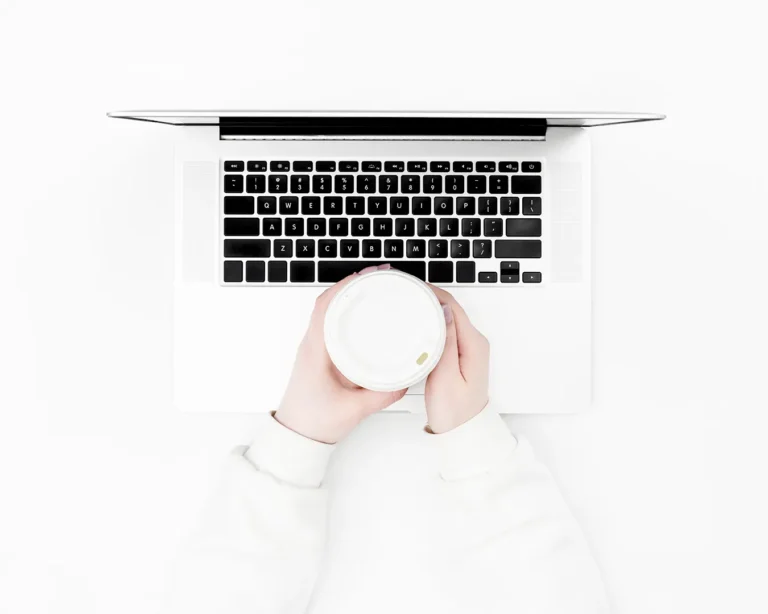 Person holding coffee cup over laptop on minimalist white workspace