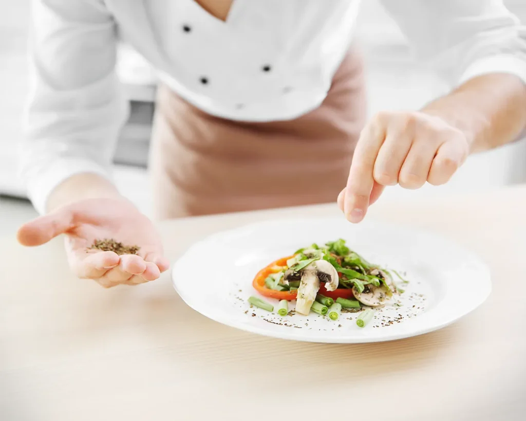 Chef carefully seasoning a plated dish in a professional kitchen, showcasing culinary craftsmanship, food quality standards, and authentic restaurant operations.