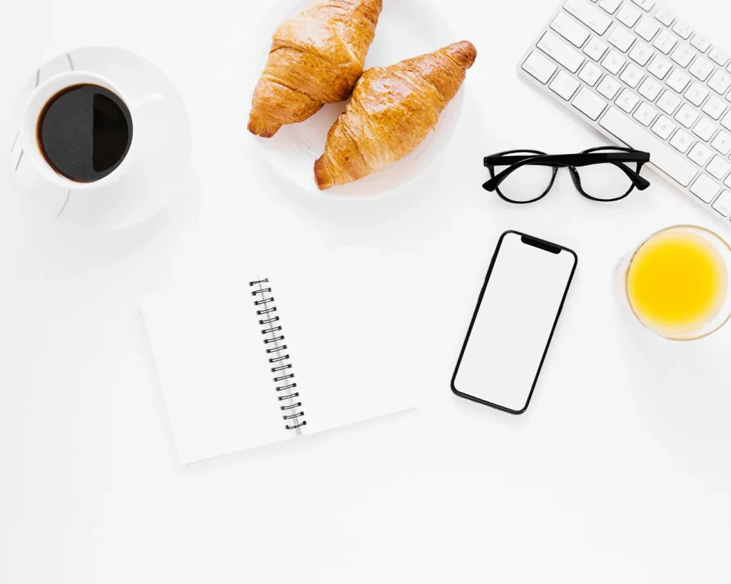 Flat lay of coffee, croissants, smartphone, notebook and keyboard on white desk
