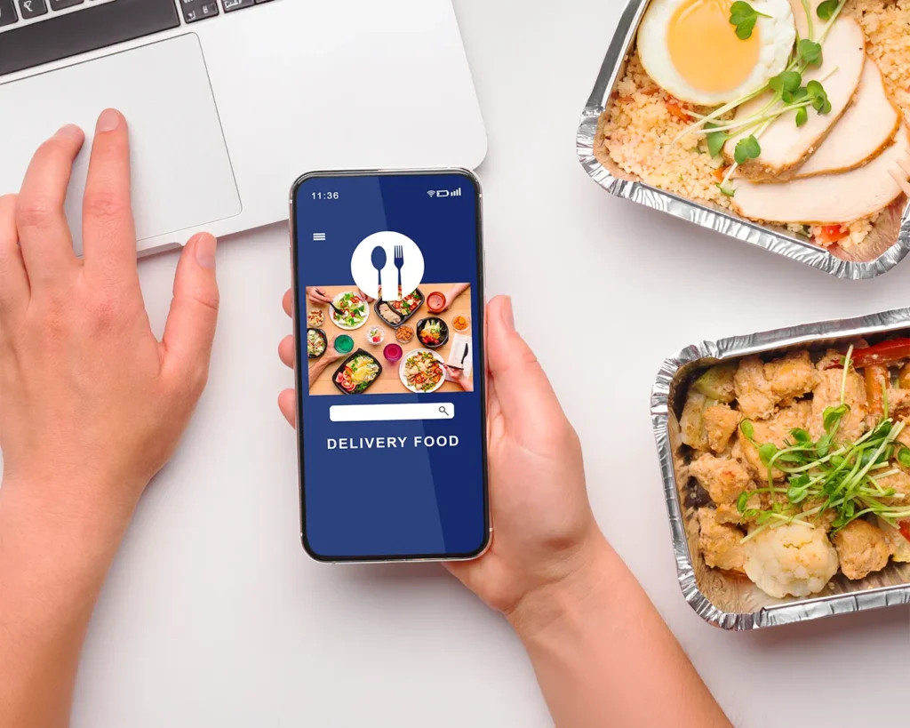 Customer using food delivery app on smartphone beside takeaway meals and laptop for online food ordering