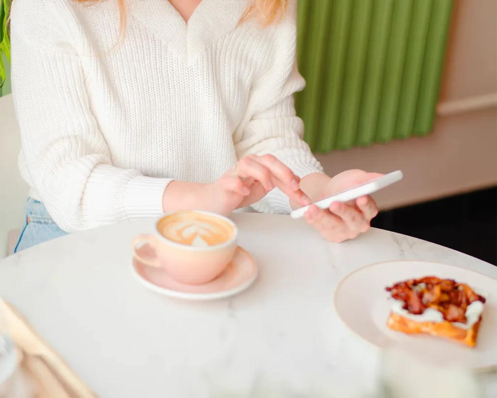 Customer using a smartphone at a café with coffee and pastry, representing mobile restaurant website browsing, online ordering, and seamless digital dining experience.