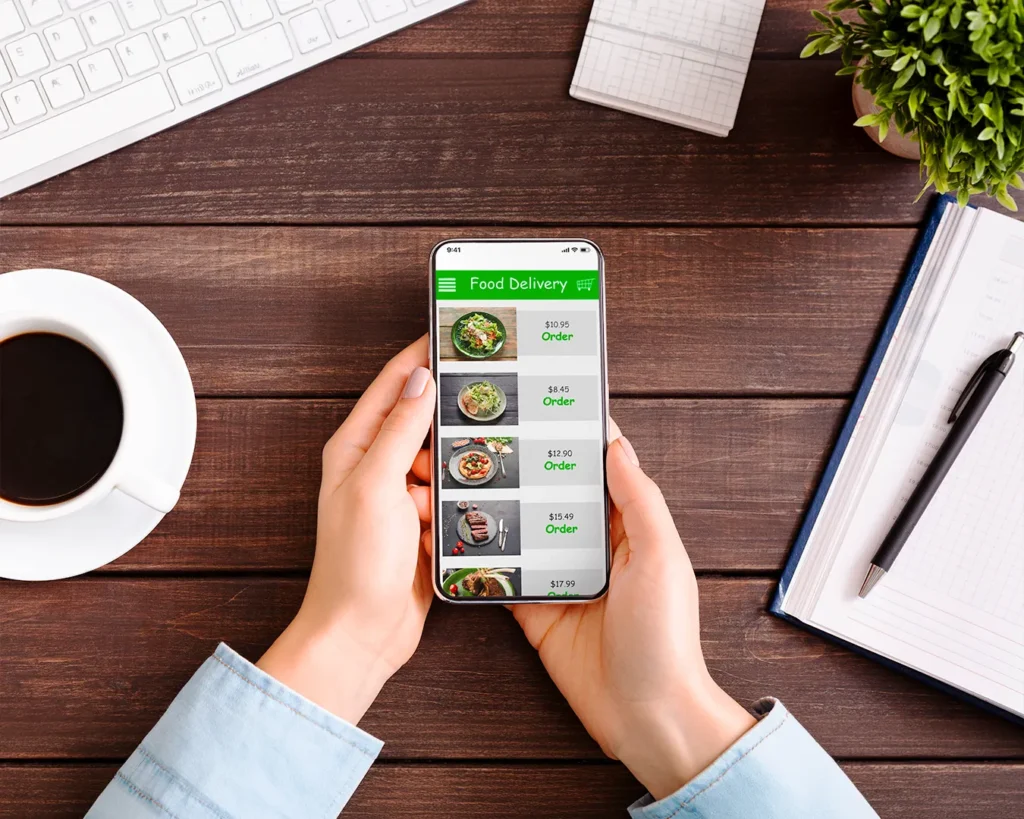 Professional ordering food delivery on smartphone at office desk with coffee and laptop workspace