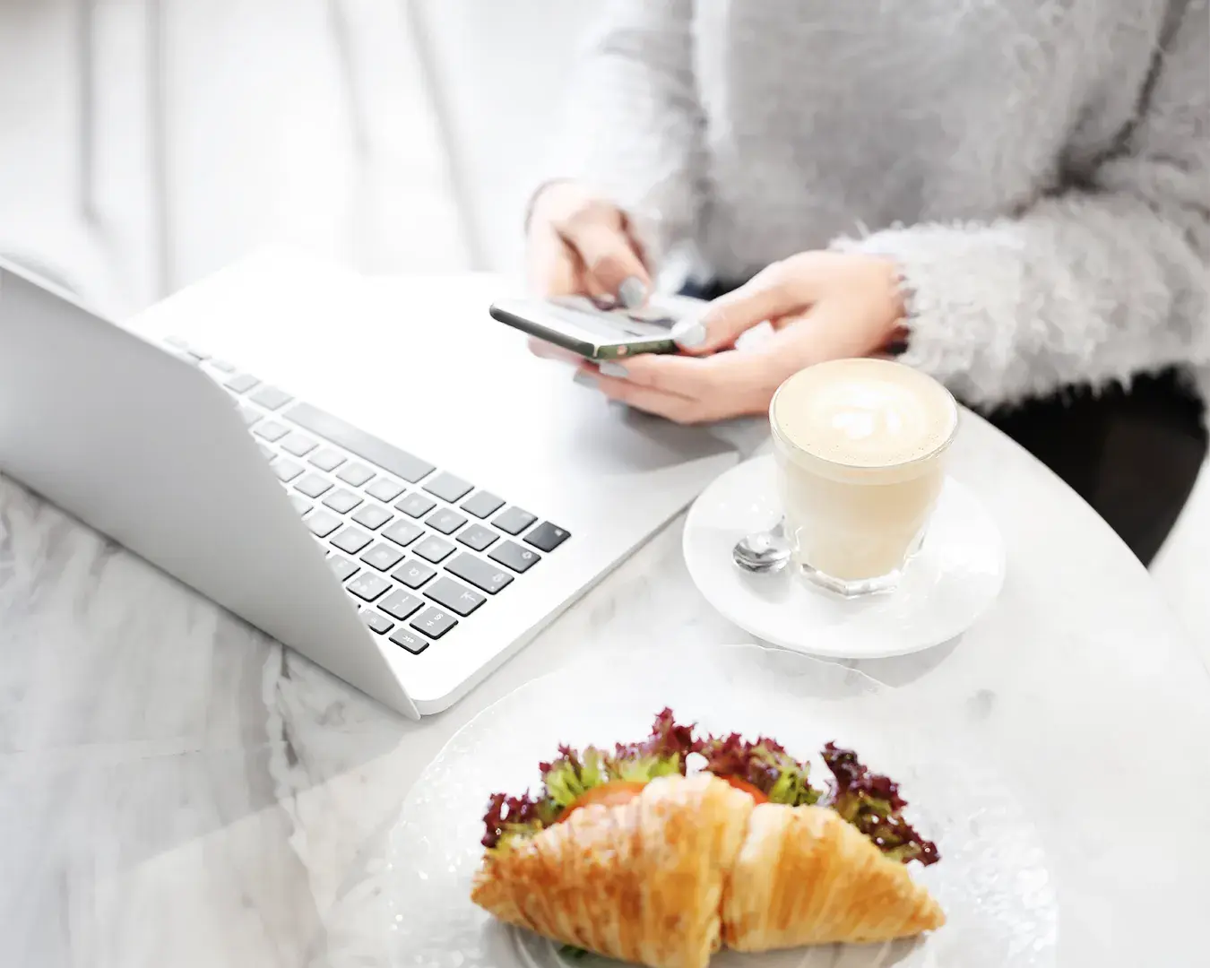 Restaurant customer browsing food delivery options on smartphone beside laptop and coffee in minimalist café setting, illustrating digital food delivery marketing strategy and online ordering behavior.